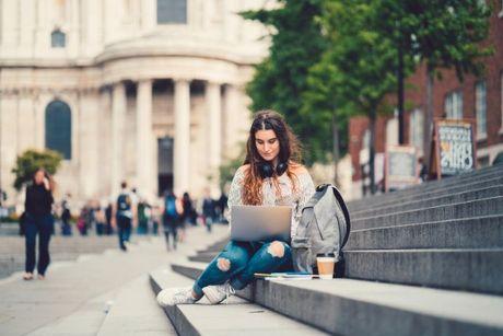 Young woman sitting on the steps of a building in a European city, applying for pre-settled status on her laptop.