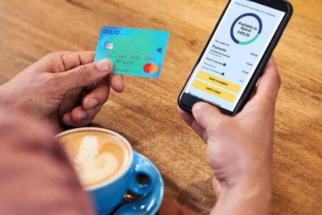 An image of a man in a coffee shop, holding his aqua credit card and looking at his phone showing the Aqua app on the screen.