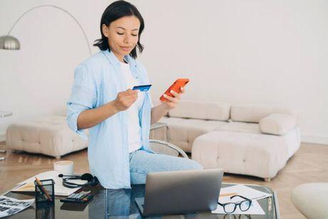 A woman in her living room with a laptop and a phone on her desk, searching about cards that have a bolt-on credit builder facility.