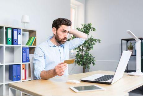 A man looking worried in front of his laptop and holding his credit card, because he miss one mortgage payment